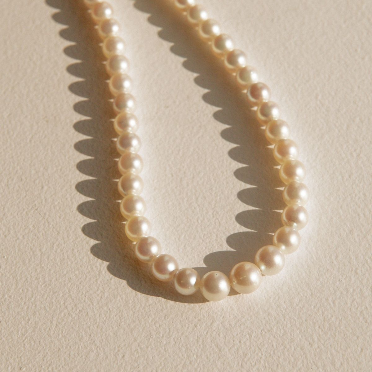 A strand of creamy white Akoya pearls arranged in a curved loop on a textured beige surface, casting soft shadows.
