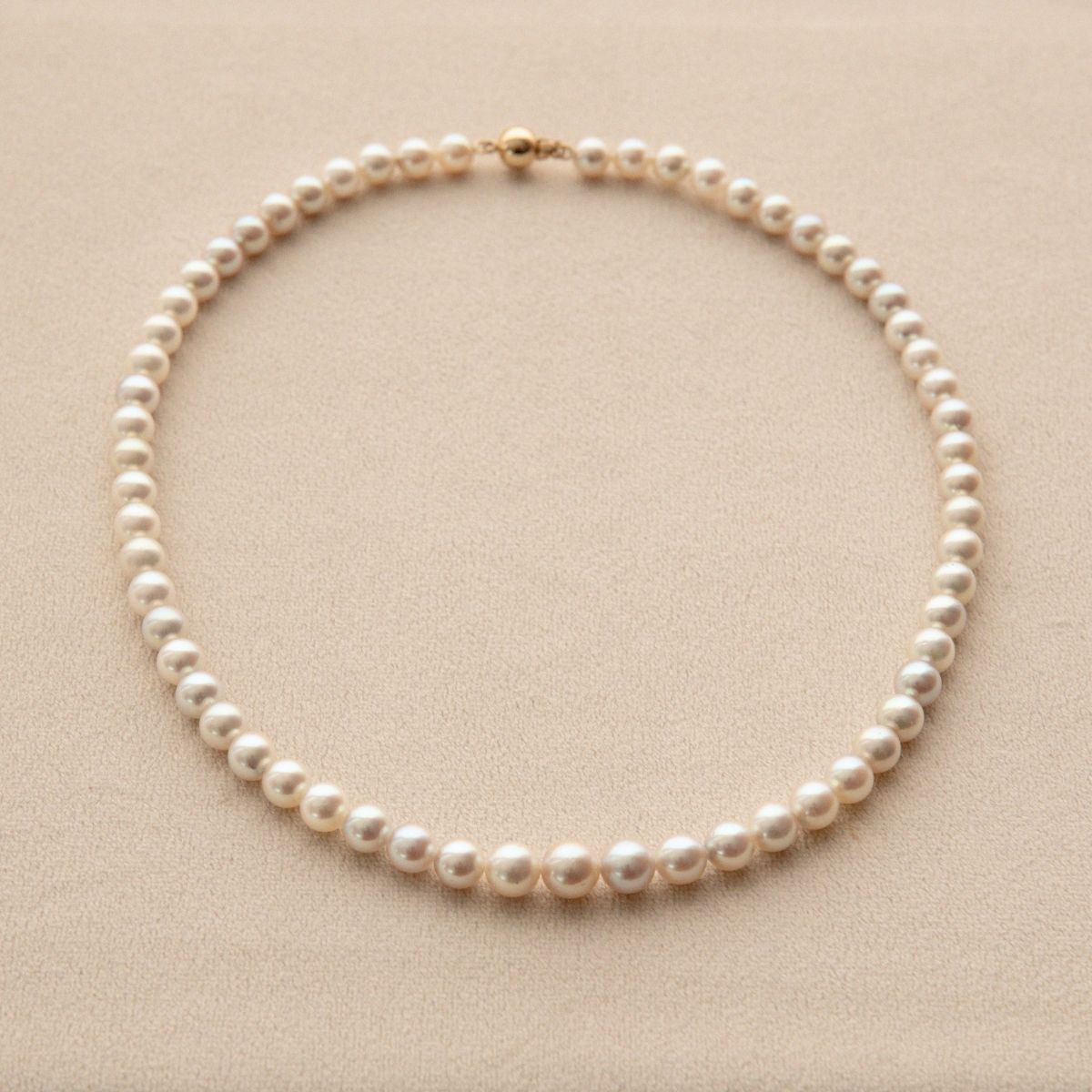 A classic Akoya pearl necklace with a gold clasp, arranged in a neat circle on a textured beige background.