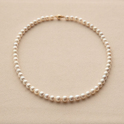 A classic Akoya pearl necklace with a gold clasp, arranged in a neat circle on a textured beige background.