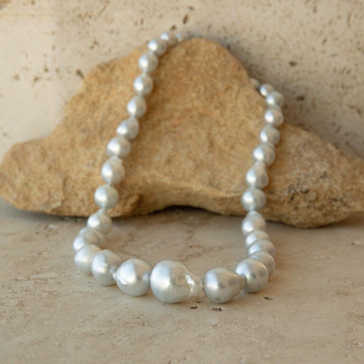 A strand necklace of irregular baroque Australian South Sea pearls draped over a tan stone on a textured beige surface.