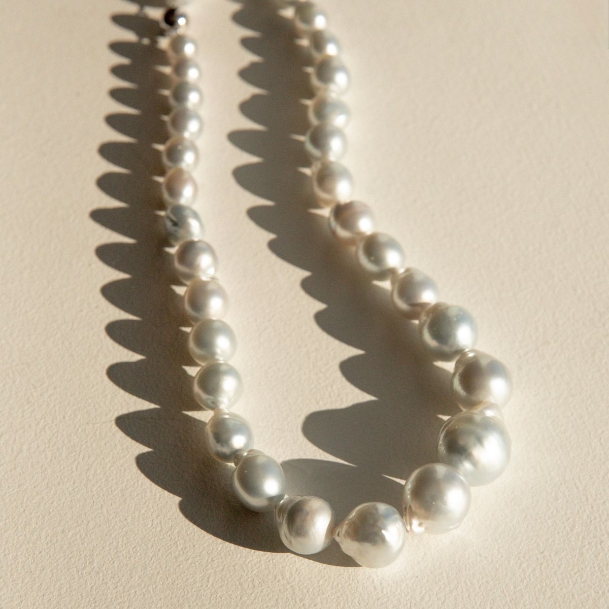 A strand of irregular Australian South Sea pearls draped in a curve shape on a textured beige surface, casting a sharp shadow to the left.