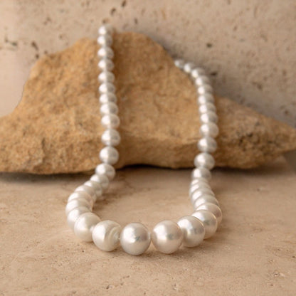 A strand of white silver toned Australian South Sea pearls draped over a natural tan rock on a matching sandy surface.
