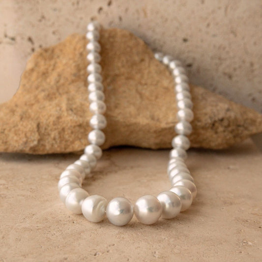 A strand of white silver toned Australian South Sea pearls draped over a natural tan rock on a matching sandy surface.