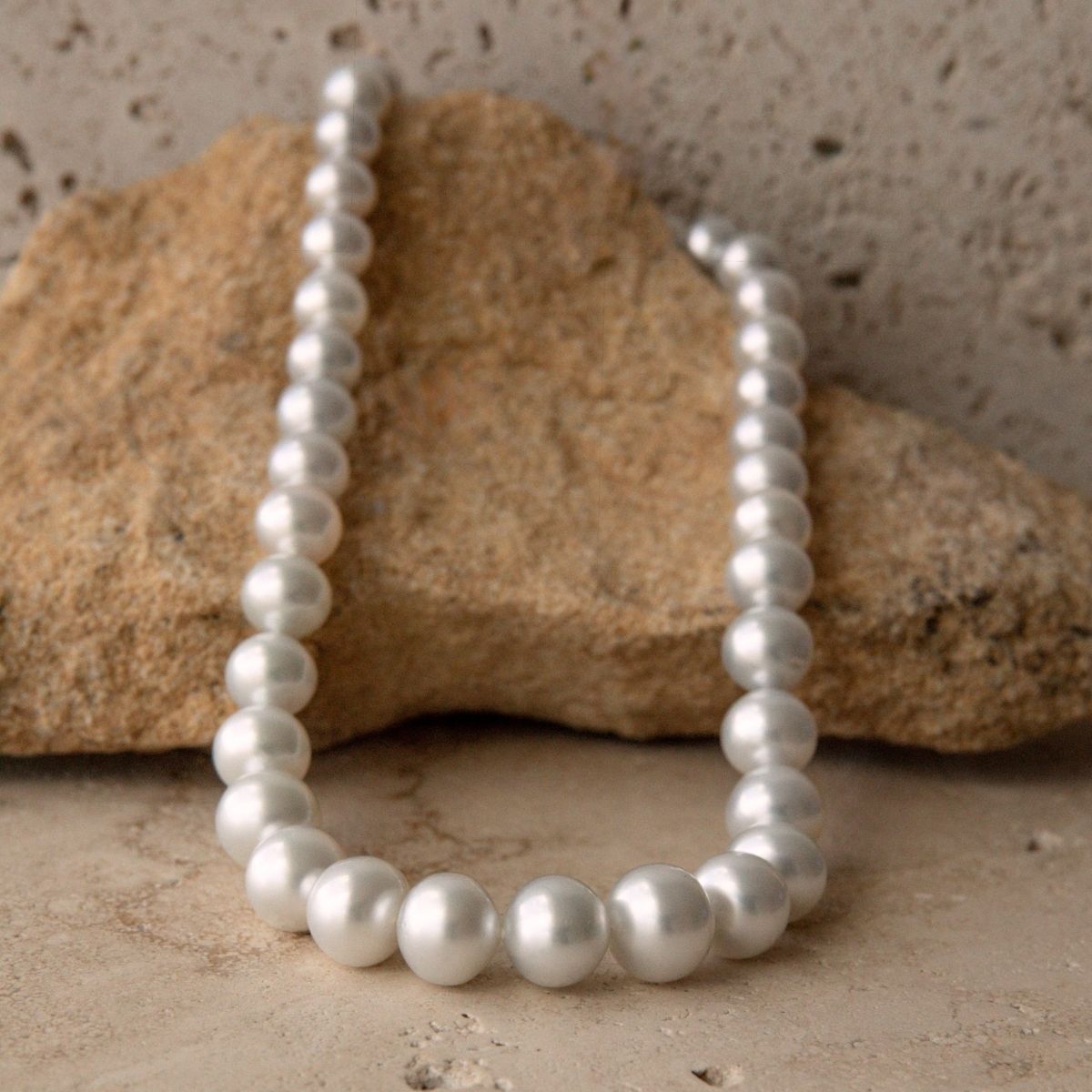 A strand necklace of white South Sea pearls arranged in a loop, resting against a natural tan rock on a textured beige stone surface.