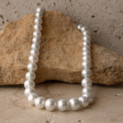 A strand necklace of white South Sea pearls arranged in a loop, resting against a natural tan rock on a textured beige stone surface.