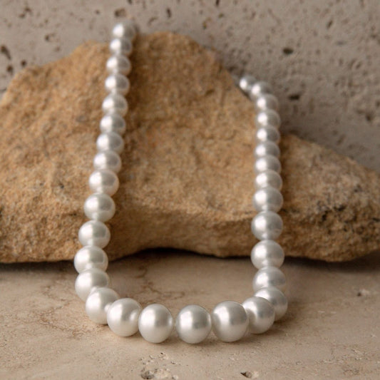 A strand necklace of white South Sea pearls arranged in a loop, resting against a natural tan rock on a textured beige stone surface.