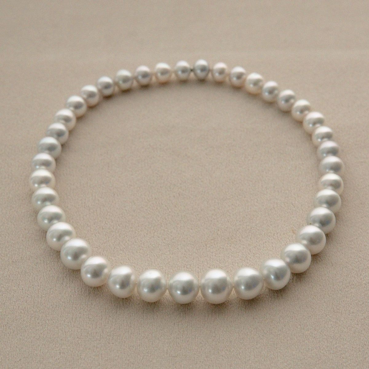 A single strand of round silver Cygnet Bay grown Australian South Sea pearls arranged in a circle on a textured beige surface.