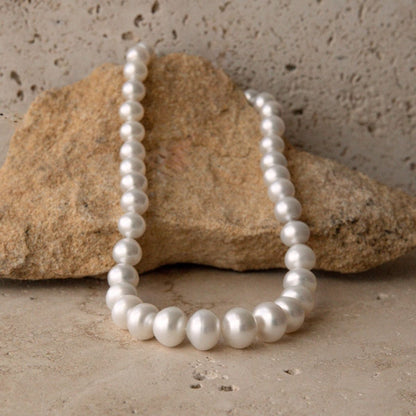 A strand of white South Sea pearls draped over a natural tan rock, set against a textured beige stone background.