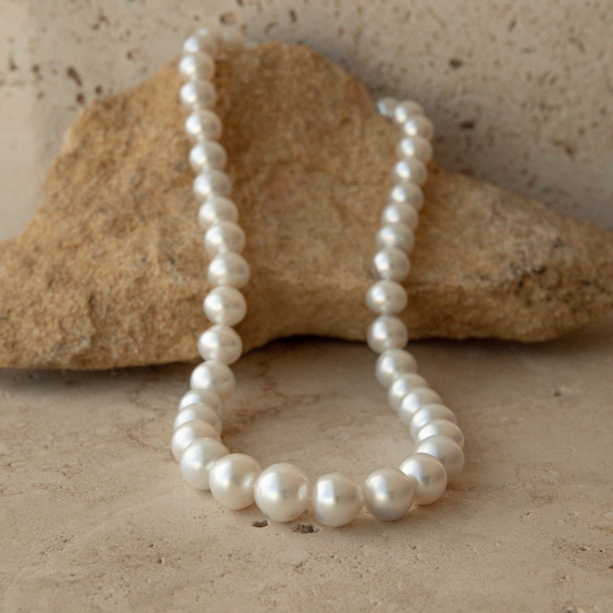 A pearl strand necklace of white button shaped Australian South Sea pearls draped over a tan rock on a textured stone surface.