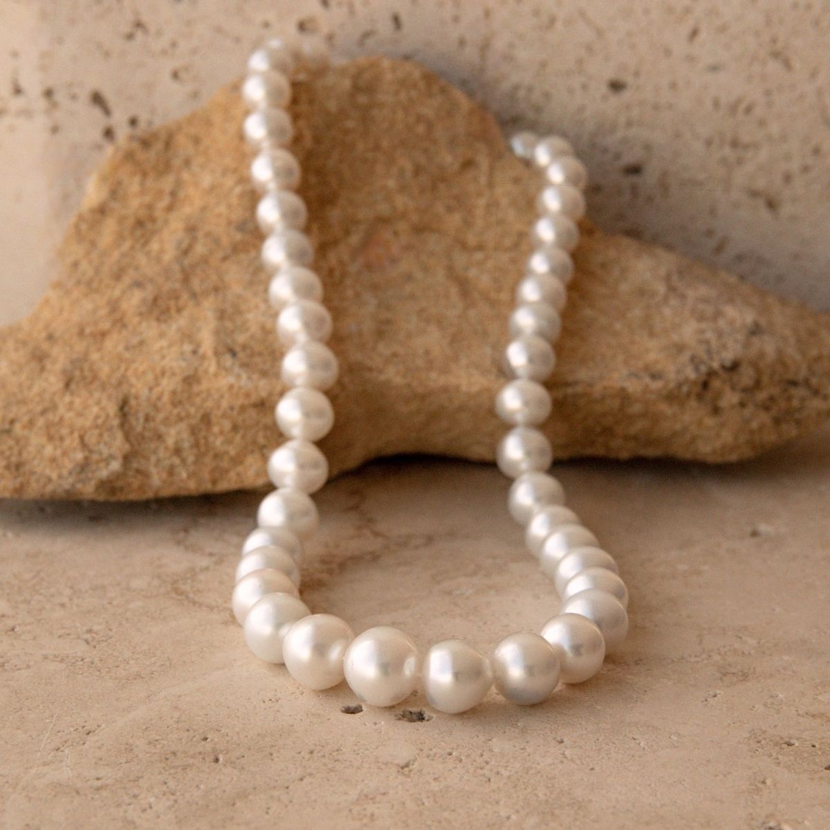 A strand of white Australian South Sea pearls draped over a natural tan rock on a textured stone surface.