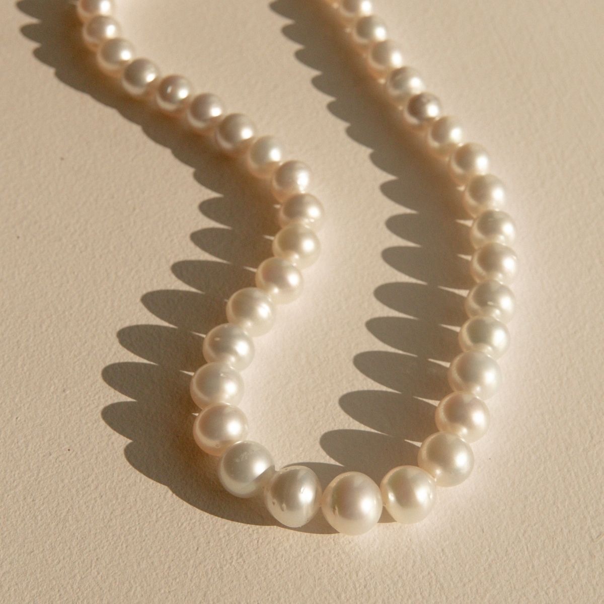 A strand of white Australian South Sea pearls arranged in a loop on a textured beige surface, casting soft shadows.