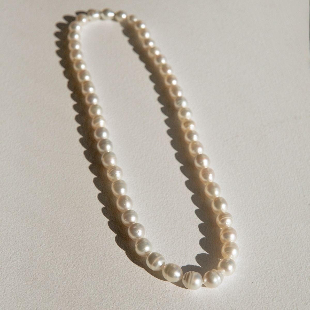 A strand of Australian South Sea pearls arranged in an oval shape on a textured off-white surface, casting soft shadows.