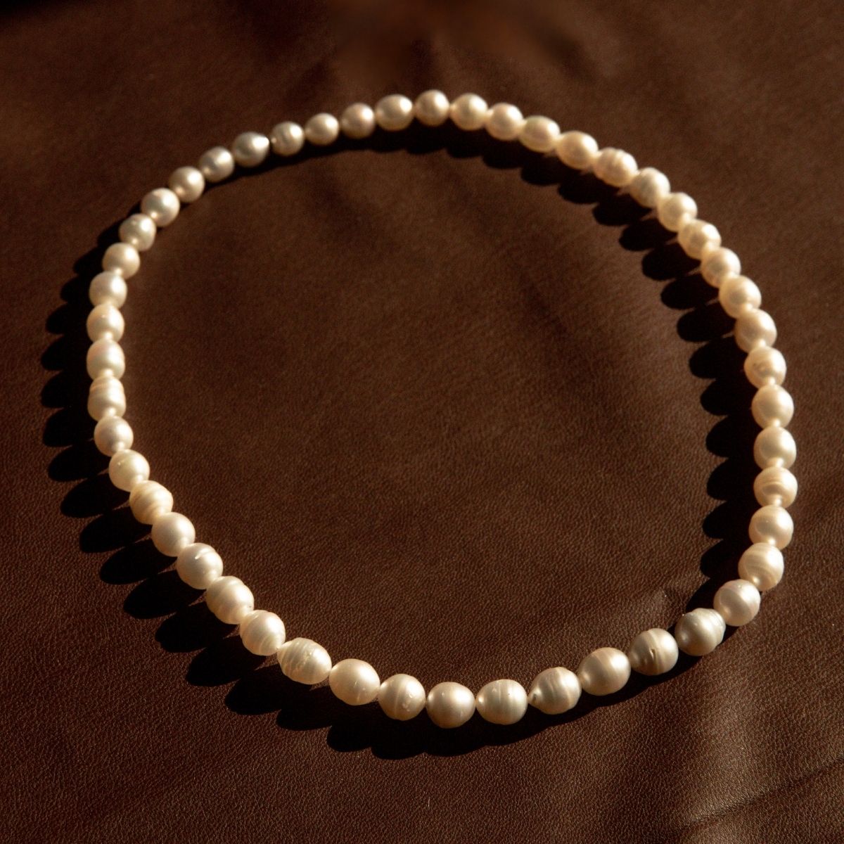 A strand of Cygnet Bay grown Australian South Sea pearls arranged in a circle on a dark brown textured fabric background.