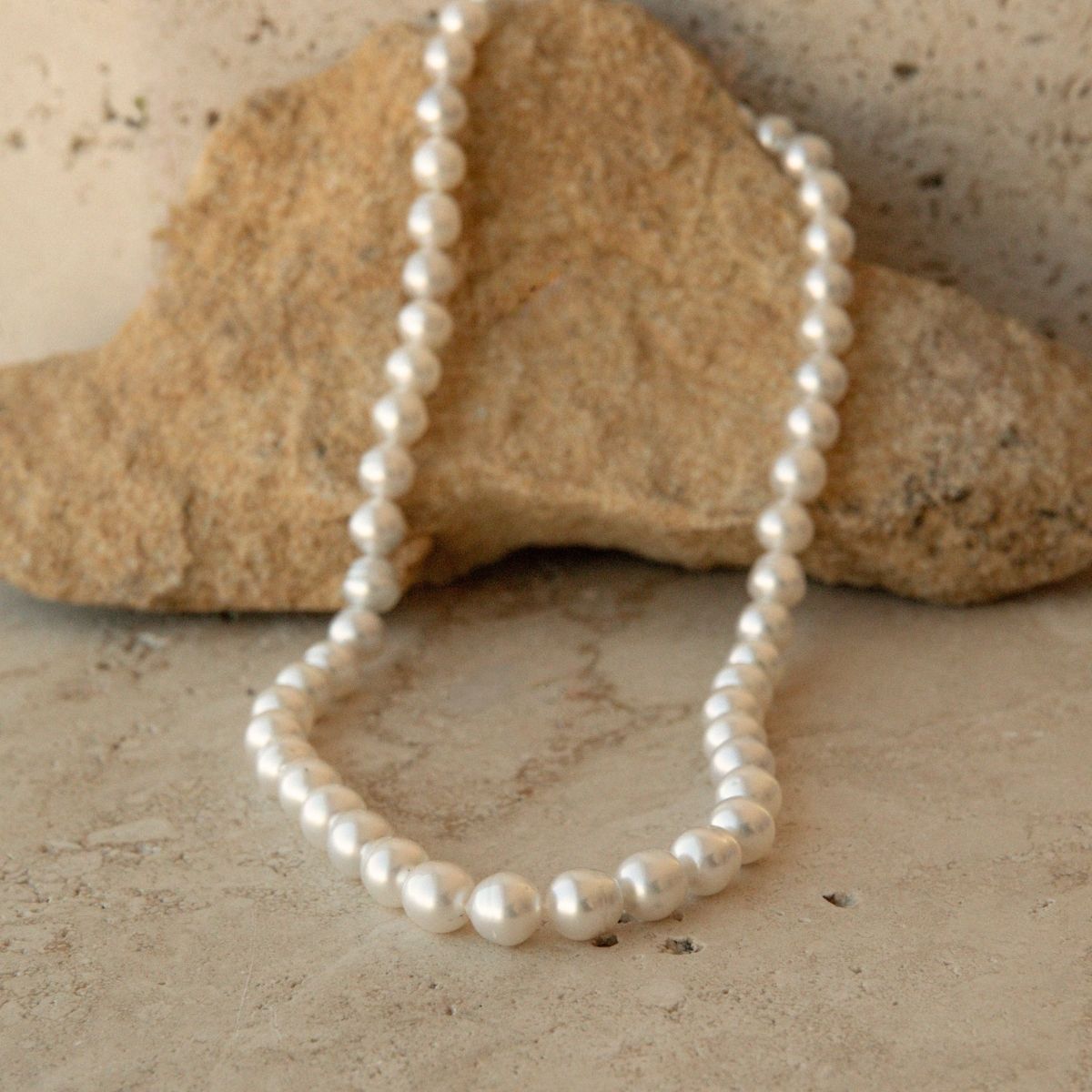 A strand of Australian South Sea pearls set in a beautifully strung pearl strand necklace, draped over a natural tan rock on a textured stone surface.
