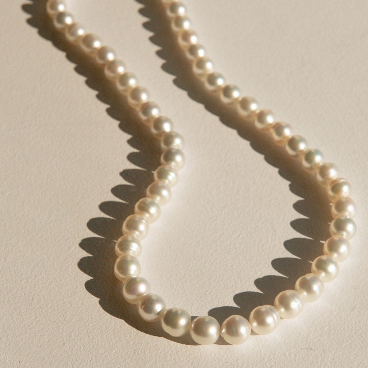 A strand of circle shaped Australian South Sea pearls arranged in a loose loop on a warm beige textured surface, casting soft shadows.