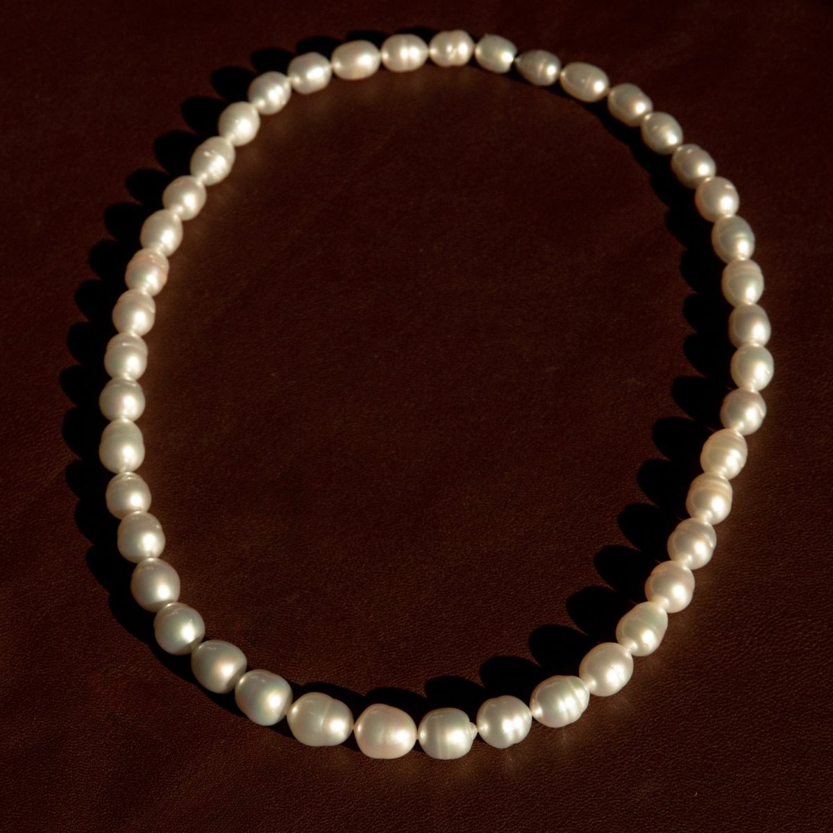 A strand of silver Australian South Sea pearls arranged in an oval shape on a dark brown textured background.