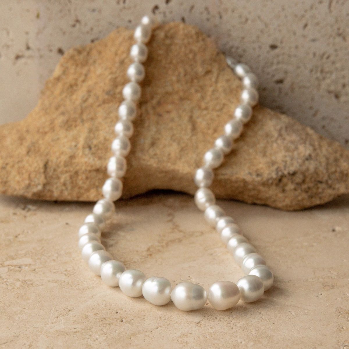A strand of silver Australian South Sea pearls draped over a natural tan rock against a textured beige stone background.