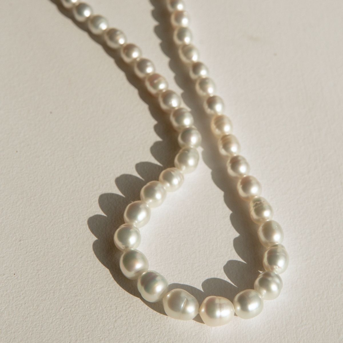 A strand of Australian South Sea pearls arranged in a loose loop on a textured off-white surface, casting soft shadows.
