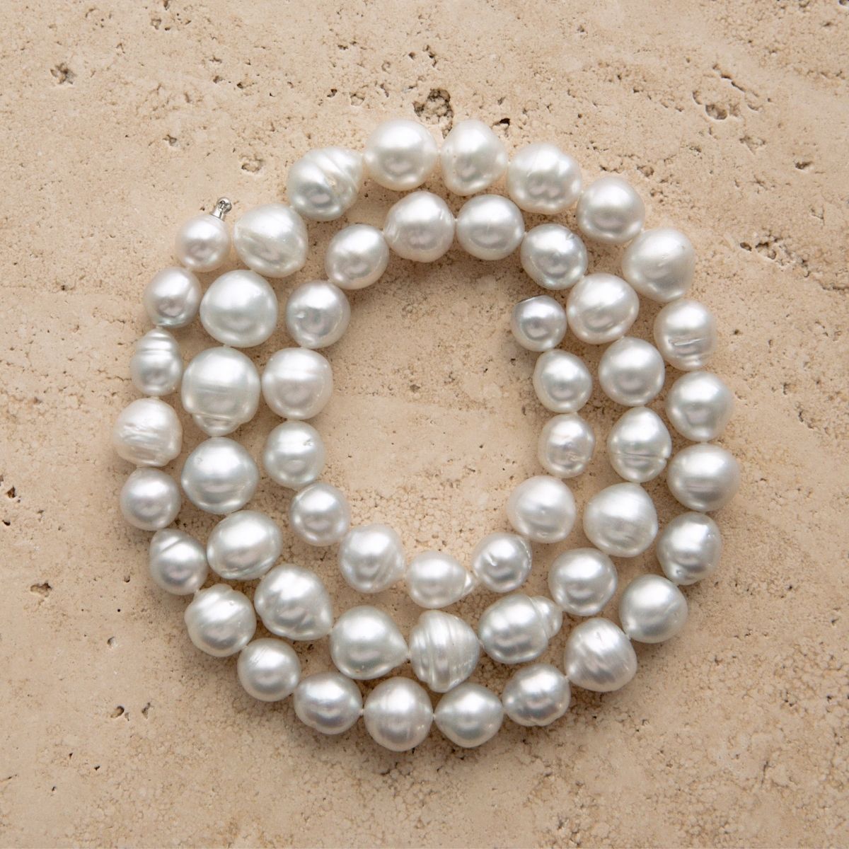 A strand of Australian South Sea pearls arranged in a loose circle on a textured beige stone surface.