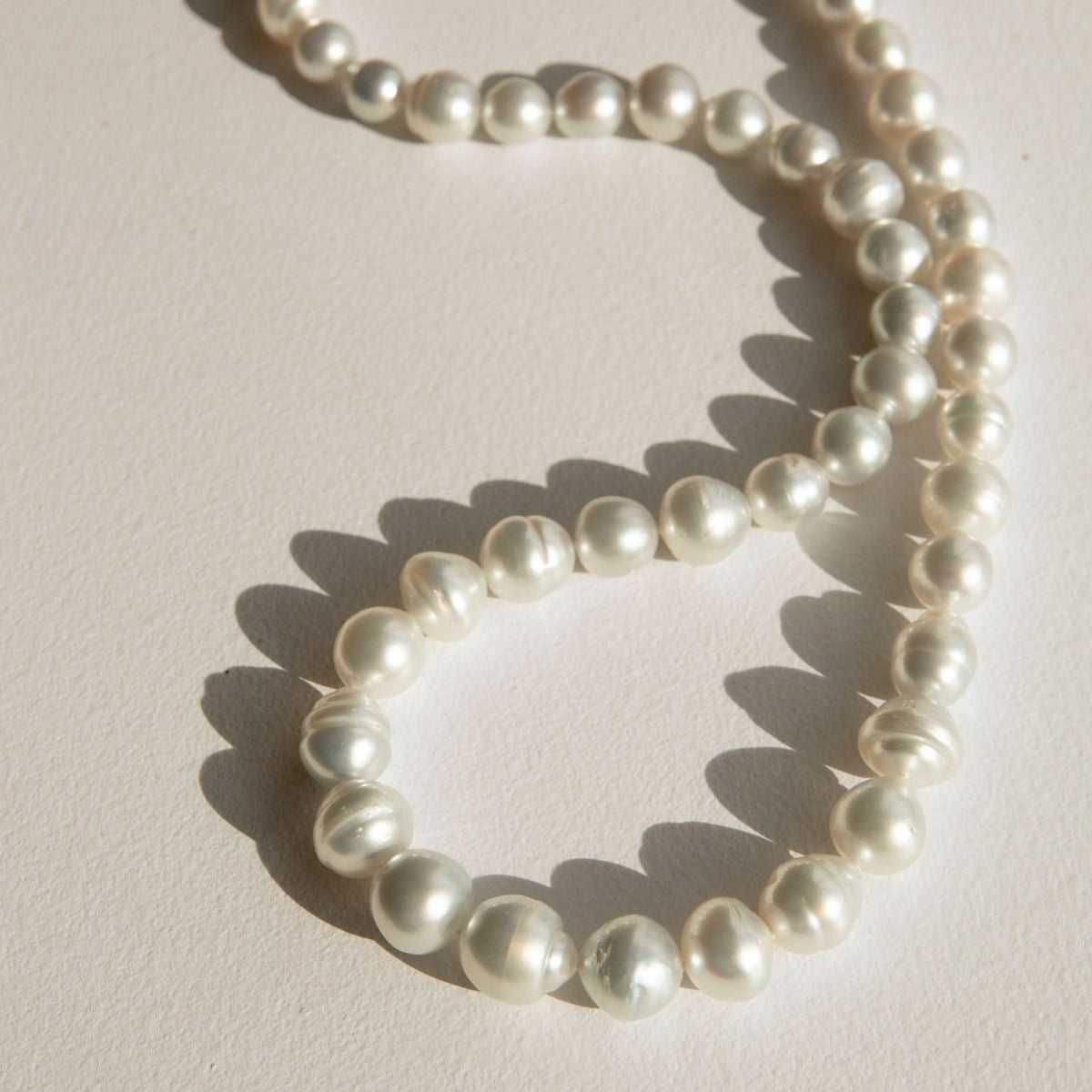 A strand of Australian South Sea pearls arranged in a loose loop on a light beige textured surface, casting soft shadows.