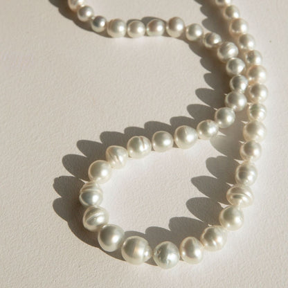 A strand of Australian South Sea pearls arranged in a loose loop on a light beige textured surface, casting soft shadows.