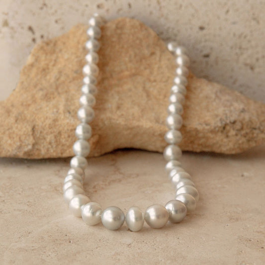 A circle pearl strand of silver Australian South Sea pearls draped over a natural tan stone on a light beige surface.