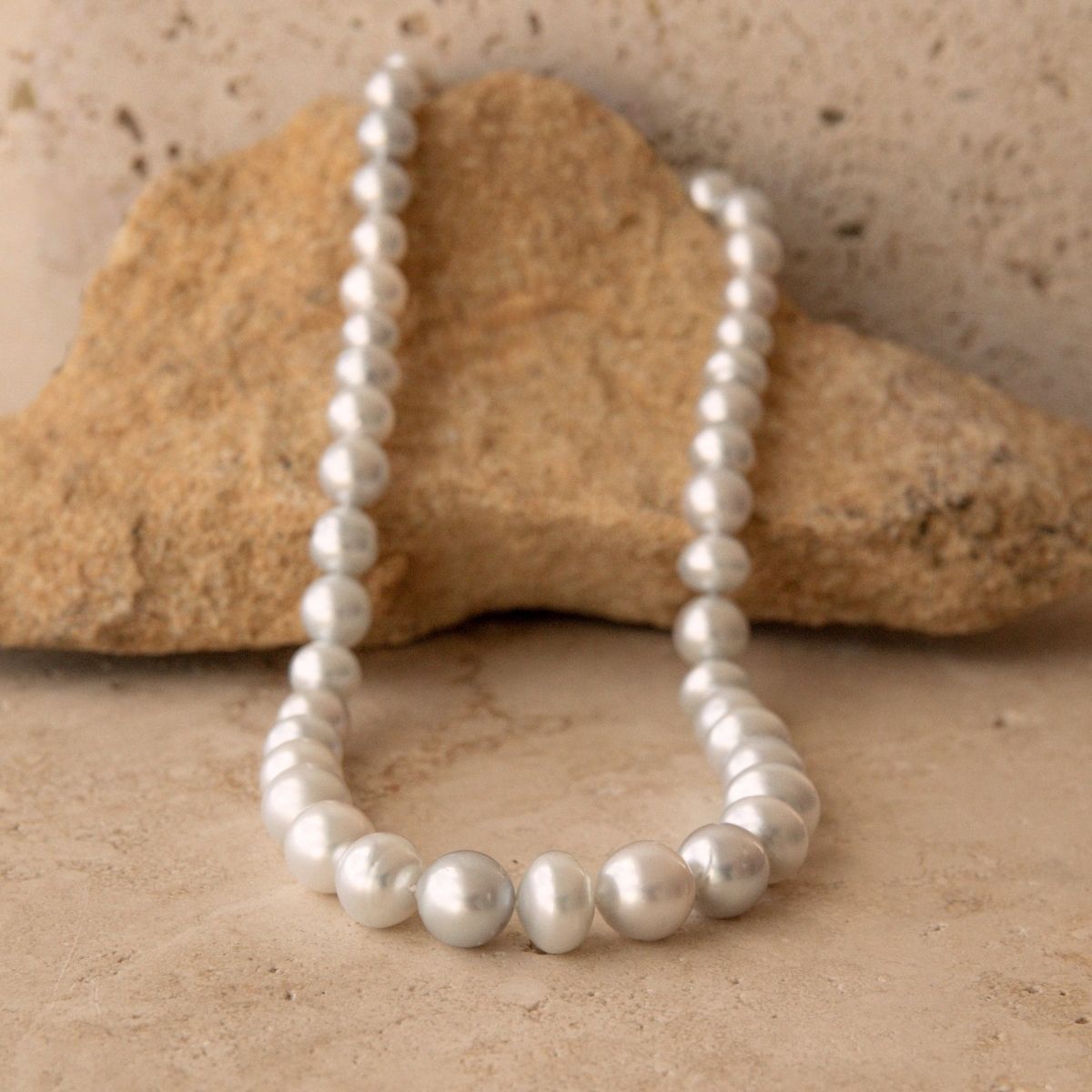 A strand of irregular silver Australian South Sea pearls draped over a natural tan rock against a textured beige stone background.