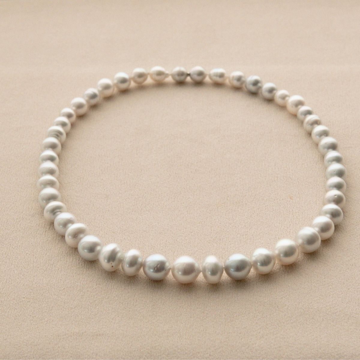 A strand of irregular silver toned Australian South Sea circle shape pearls arranged in a circle on a textured beige background.