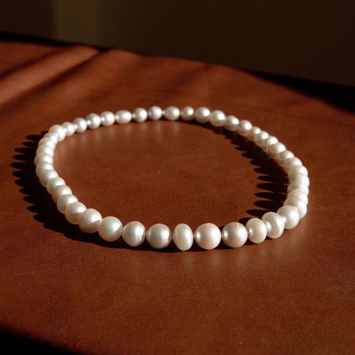 A strand necklace of white Australian South Sea pearls arranged in a circle on a brown leather surface, with dramatic warm lighting casting shadows.