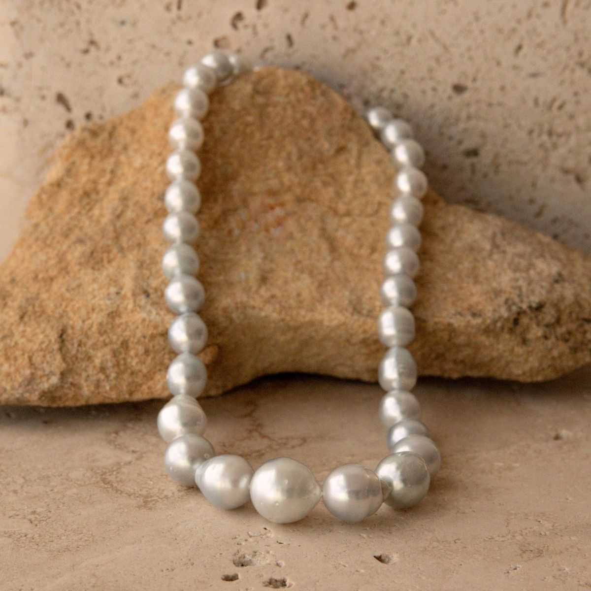 A strand of lustrous silver-blue toned Australian South Sea circle pearls draped over a tan stone on a textured beige surface.