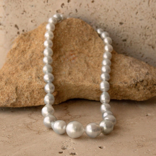 A strand of lustrous silver-blue toned Australian South Sea circle pearls draped over a tan stone on a textured beige surface.
