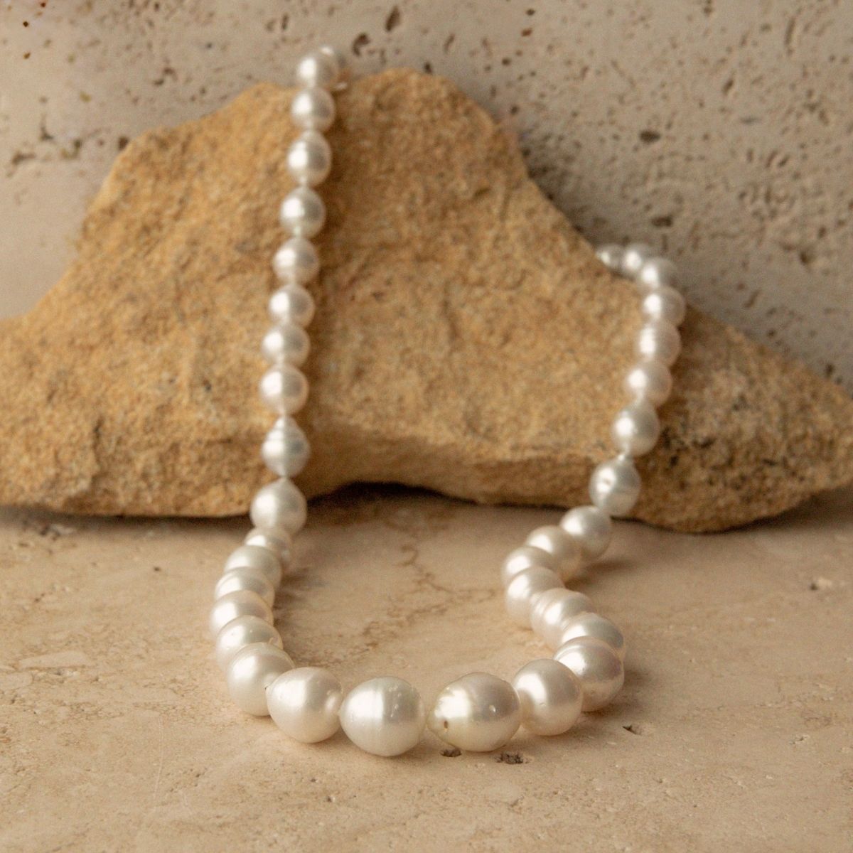 A strand of Cygent Bay grown Australian South Sea pearls draped over a tan rock against a textured beige stone background.