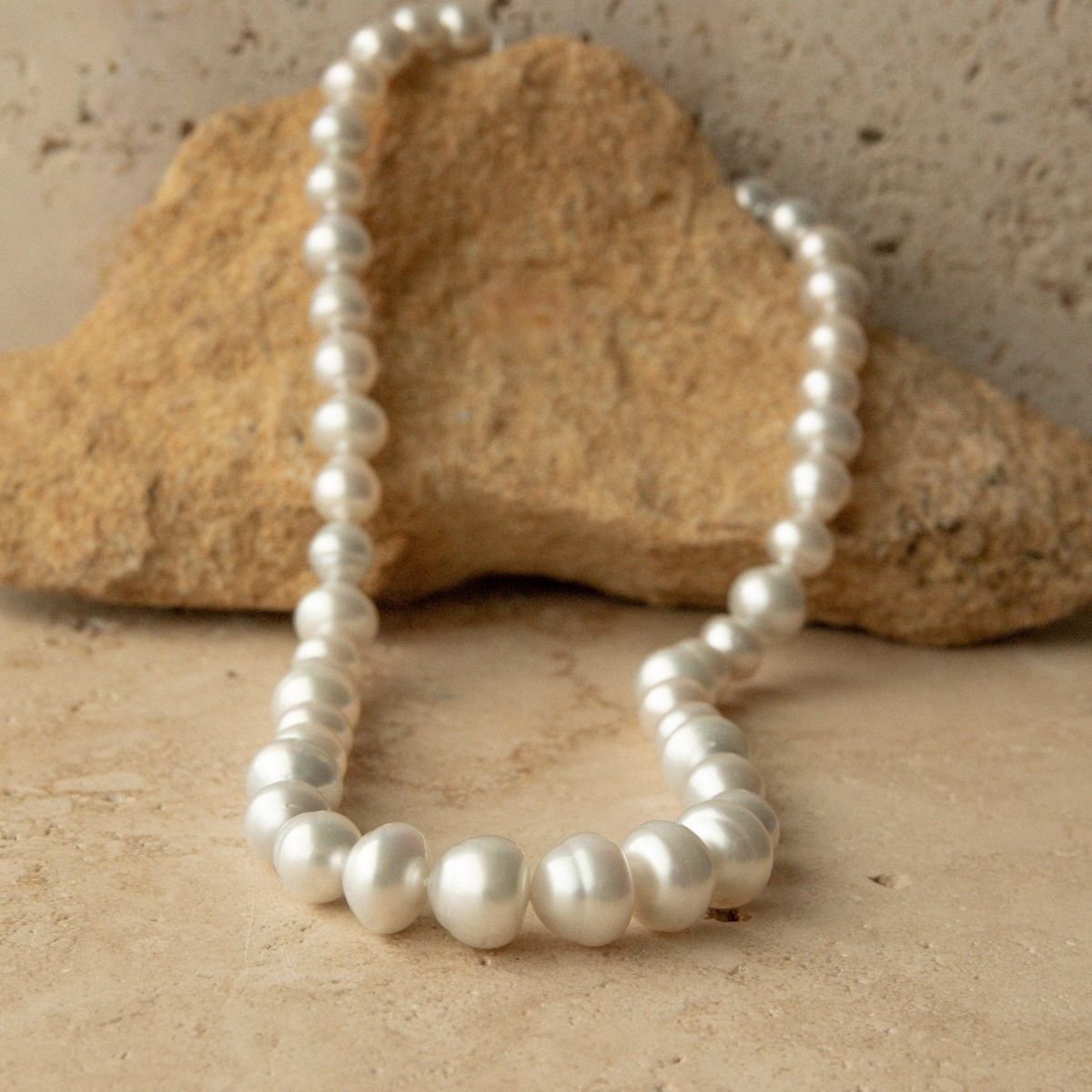 A strand of Australian South Sea pearls draped over a natural tan rock on a textured beige stone surface.