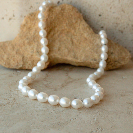 A strand of white Australian South Sea pearls arranged in a loop on a light stone surface, with a larger tan rock prop behind it.