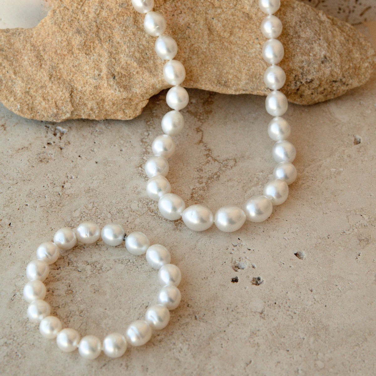 A white Australian South Sea pearl necklace and matching pearl bracelet arranged on a light stone surface with a larger tan rock prop in the background.