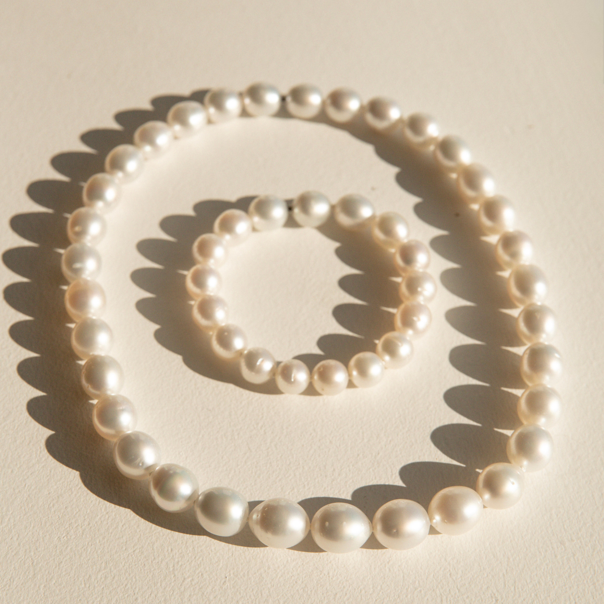 A strand of white Australia South Sea pearls from Cygnet Bay Pearl Farm in a circle on a warm cream background, with a matching bracelet nested inside. The beads are photographed in direct sunlight, casting sharp shadows.