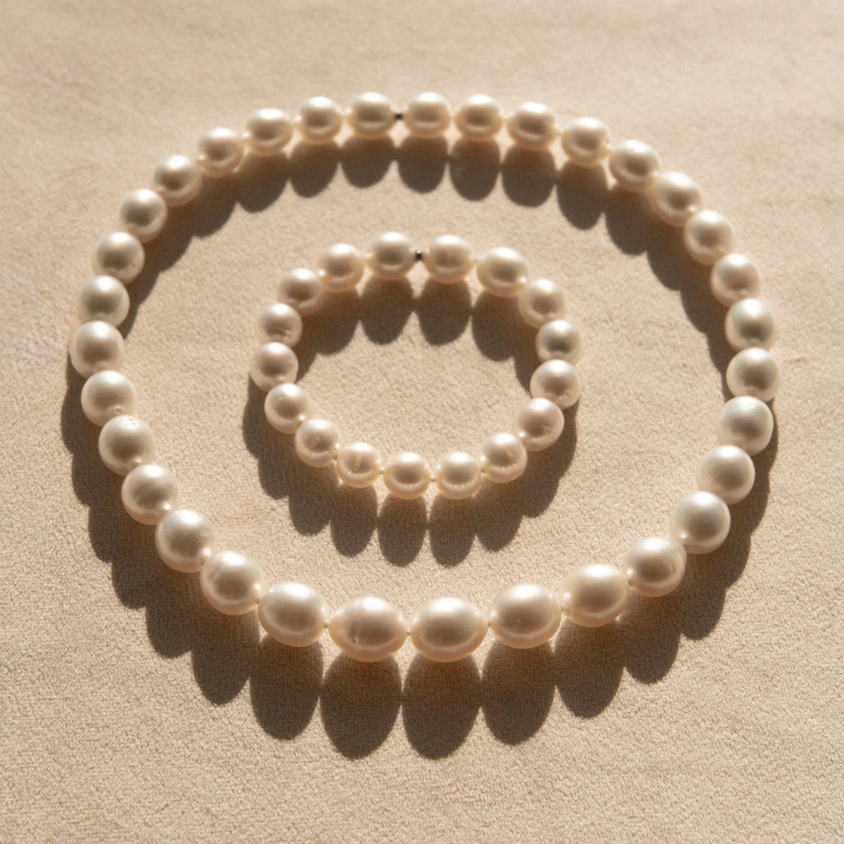 A set of matching pearl strand neclace and bracelet in graduated sizes, arranged in a circle pattern on a textured beige surface with soft shadows.