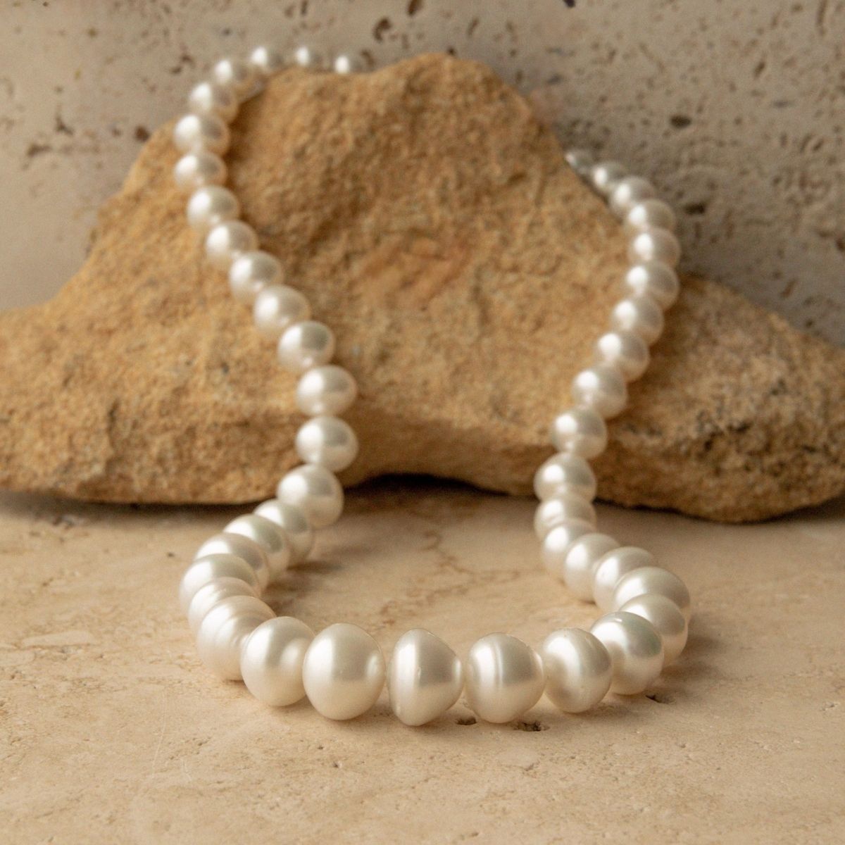 A strand of white Australian South Sea pearls arranged in a loop on a beige stone surface, with a larger tan rock prop behind them against a textured beige wall.