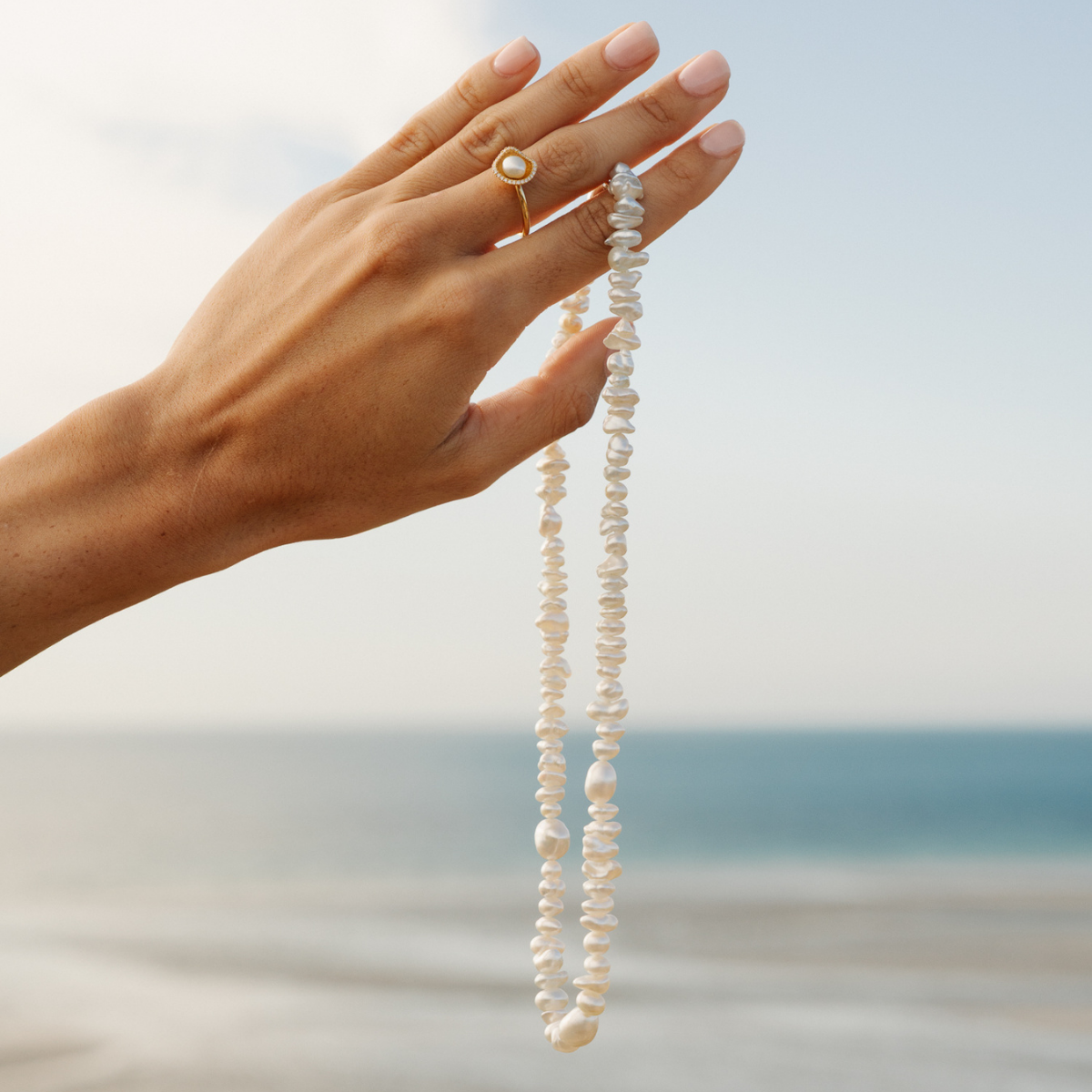 Hand holding a long keshi pearl necklace with a blurred beach background