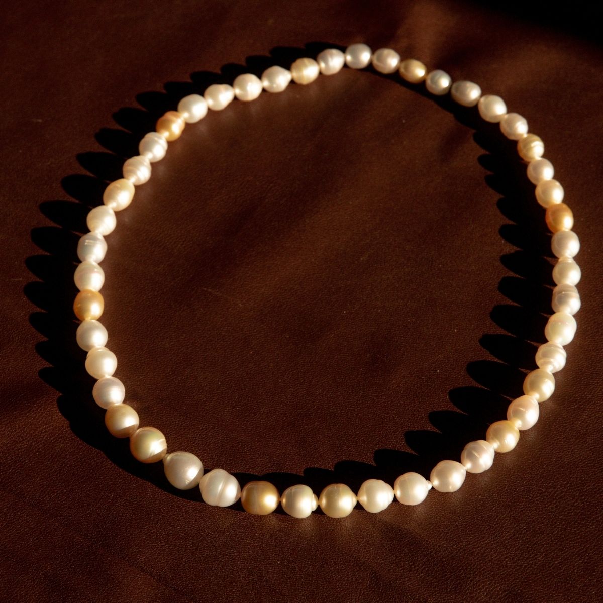 A strand of Australian South Sea white and cream pearls arranged in a circle on a dark brown textured surface, casting soft shadows.