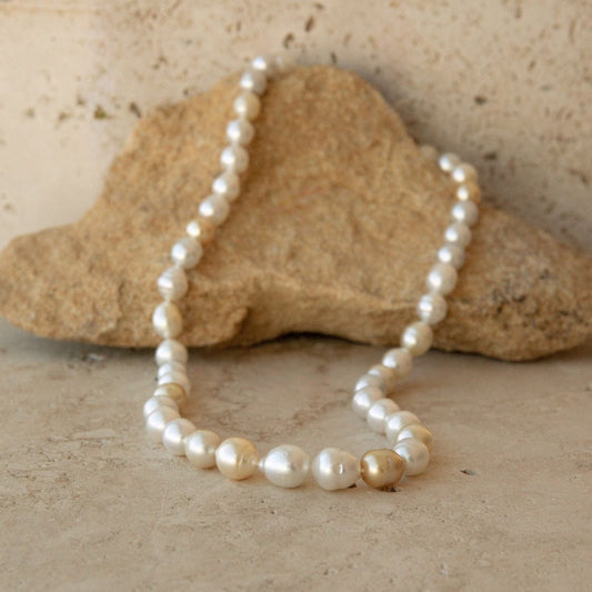 A strand of Australian South Sea pearls in warm champagne-gold tones, draped over a natural tan stone on a textured beige surface.
