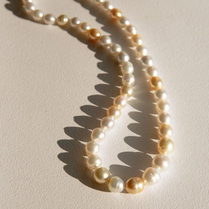 A strand of Australian South Sea pearls in cream, ivory, and light gold tones, arranged in a loose loop on a textured beige surface with sharp sunlight shadows.