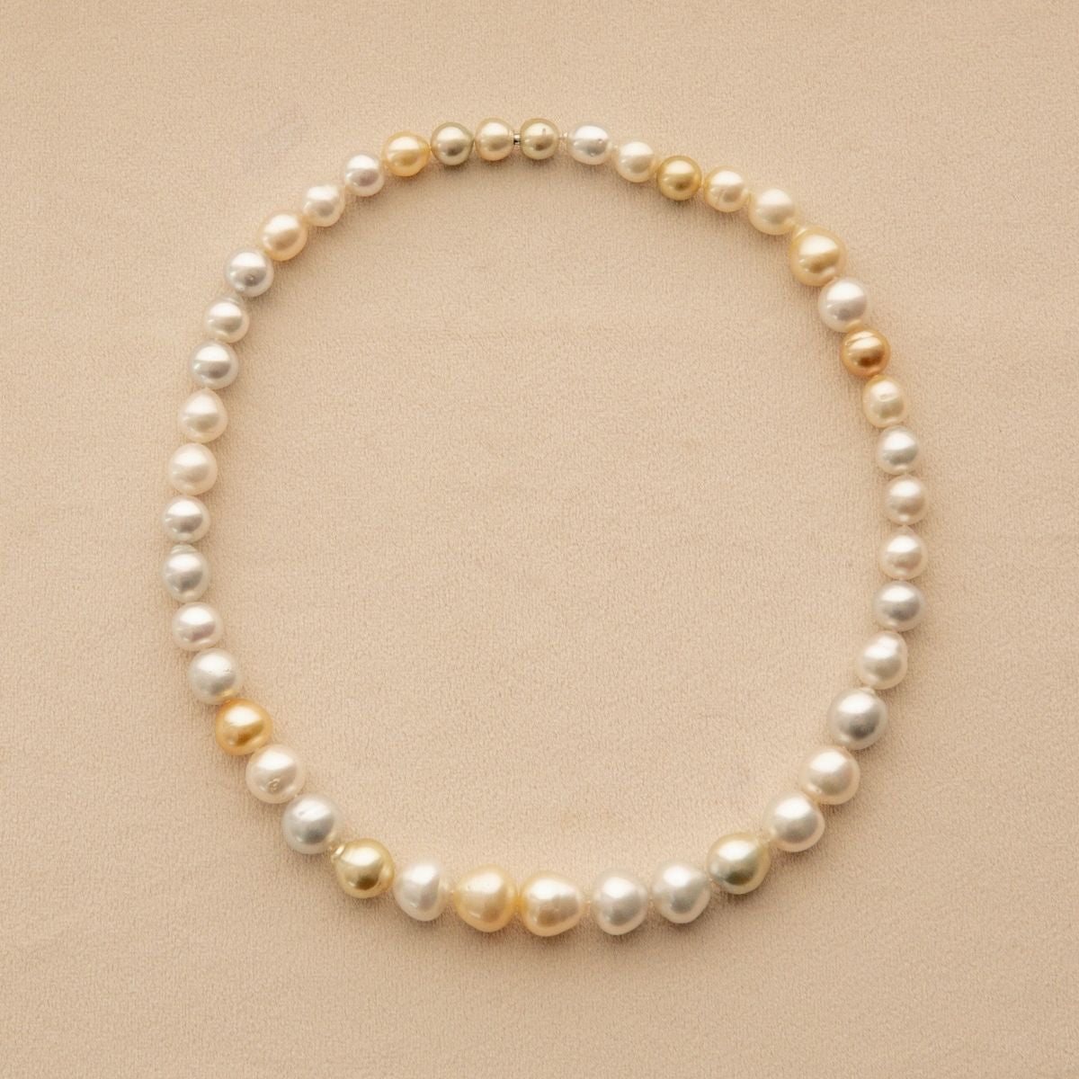 A strand of Cygnet Bay grown Australian South Sea pearls in white, cream, and soft gold, arranged in a circle on a textured beige background.