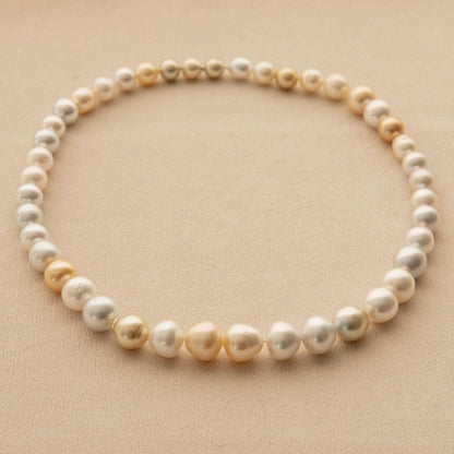 A strand necklace made of mixed shape Australian South Sea pearls in white, cream, and soft gold tones, arranged in a circle on a warm beige textured background.