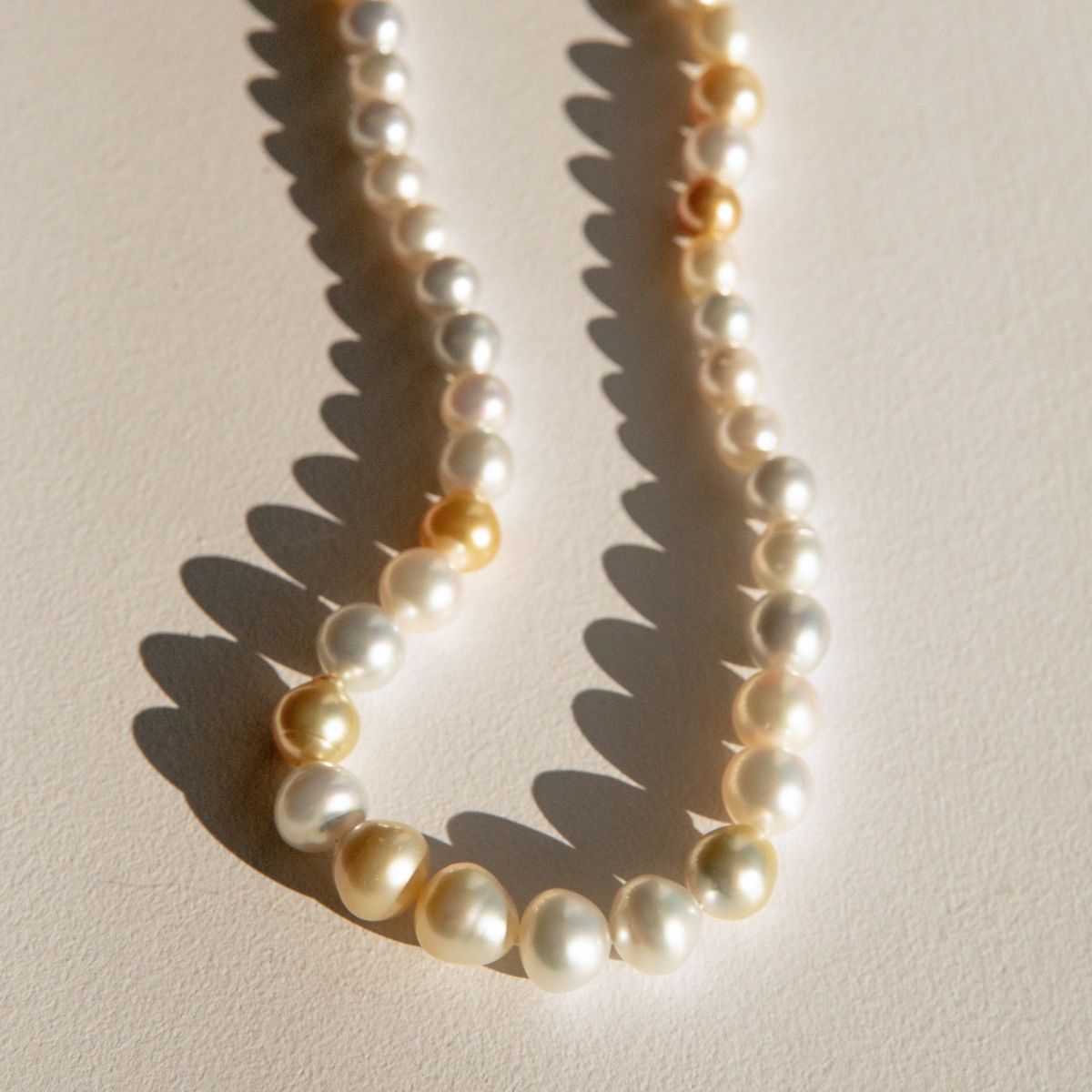 A strand of irregular shape Australian South Sea pearls in white, cream, and soft gold tones, arranged in a loose curve on a warm beige surface with sharp sunlight shadows.