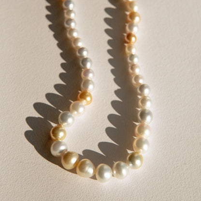 A strand of irregular shape Australian South Sea pearls in white, cream, and soft gold tones, arranged in a loose curve on a warm beige surface with sharp sunlight shadows.