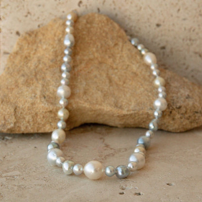A strand necklace made of white, cream, and silver Australian South Sea and Akoya pearls draped over a tan rock on a stone surface.