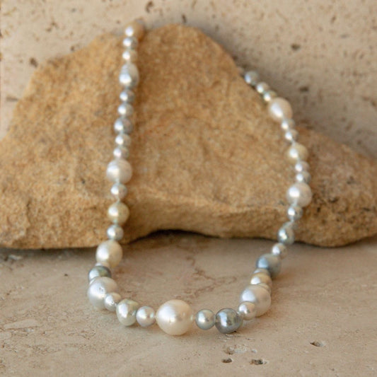 A strand necklace made of white, cream, and silver Australian South Sea and Akoya pearls draped over a tan rock on a stone surface.