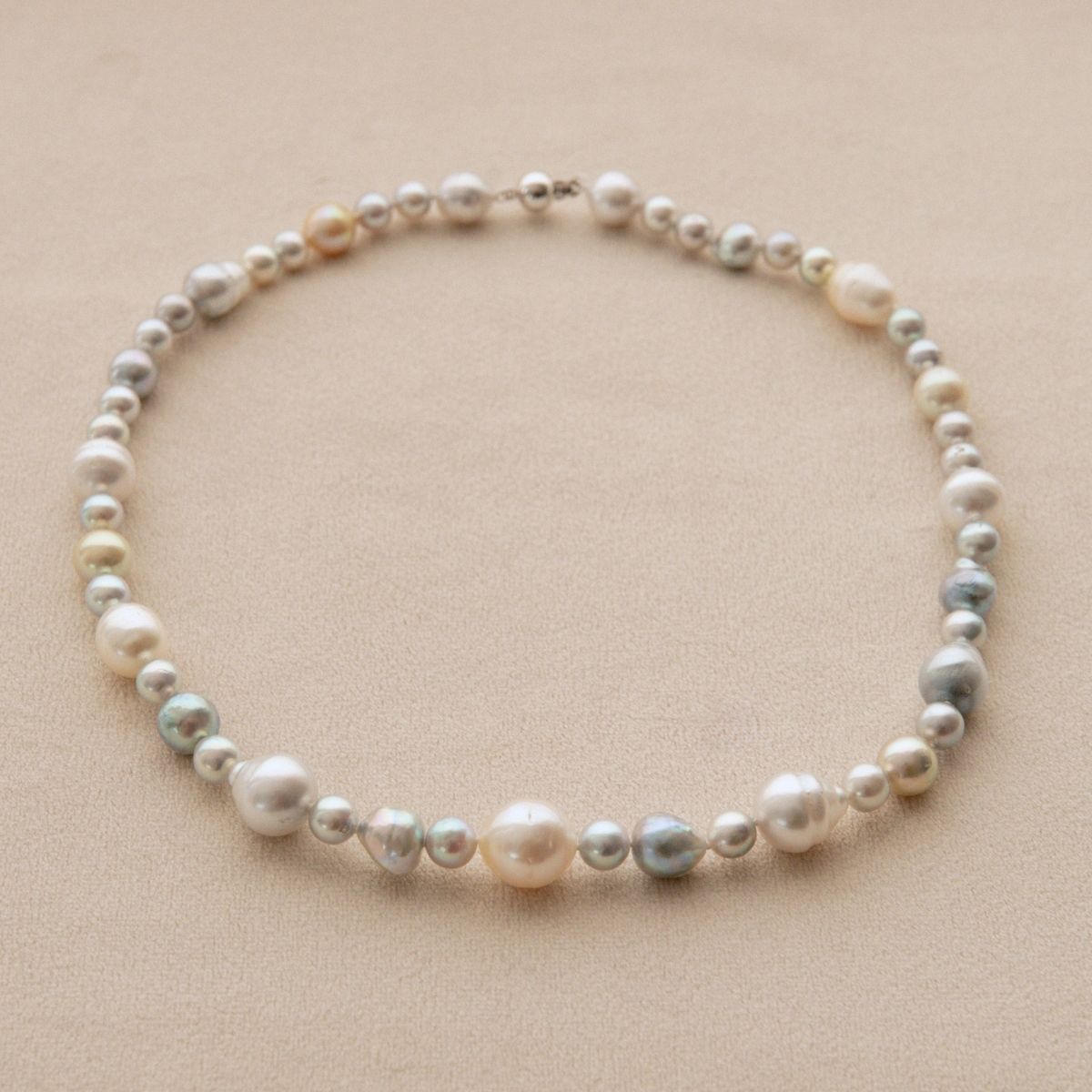 A pearl strand necklace with a mix of white, cream, and iridescent blue Australian South Sea and Akoya pearls, arranged in a circle on a neutral beige background.