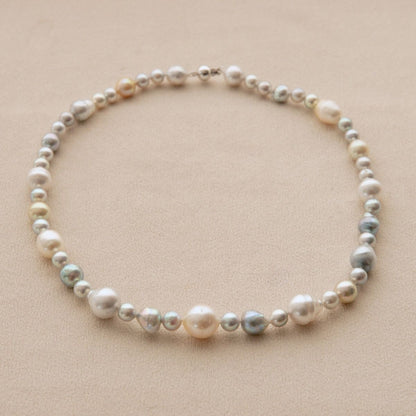 A pearl strand necklace with a mix of white, cream, and iridescent blue Australian South Sea and Akoya pearls, arranged in a circle on a neutral beige background.